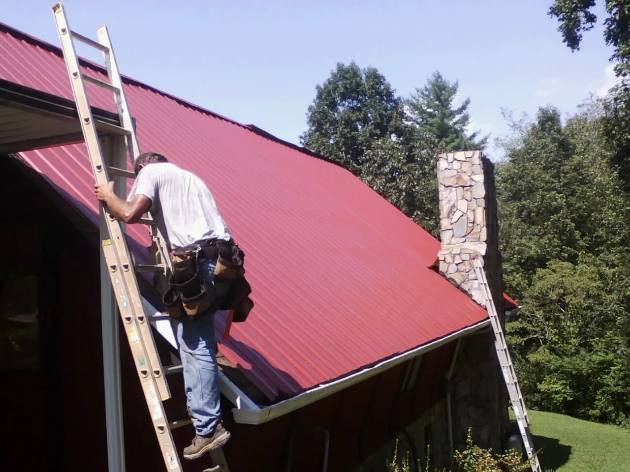 Roofer on a ladder beside a red metal roof and stone chimney — Soffit & Fascia Repair by Roofing Brothers in North Truro